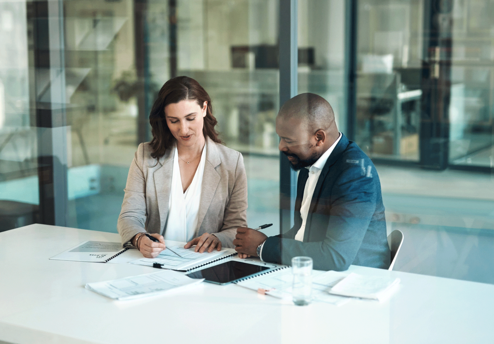 Two people in office setting, in suits, sitting at desk looking over notes in a notebook