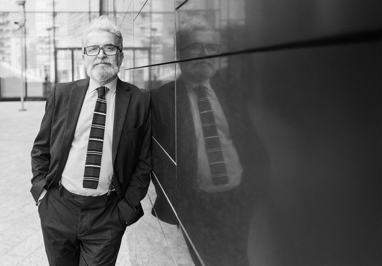 Senior businessman with hands in pockets leaning outside office building