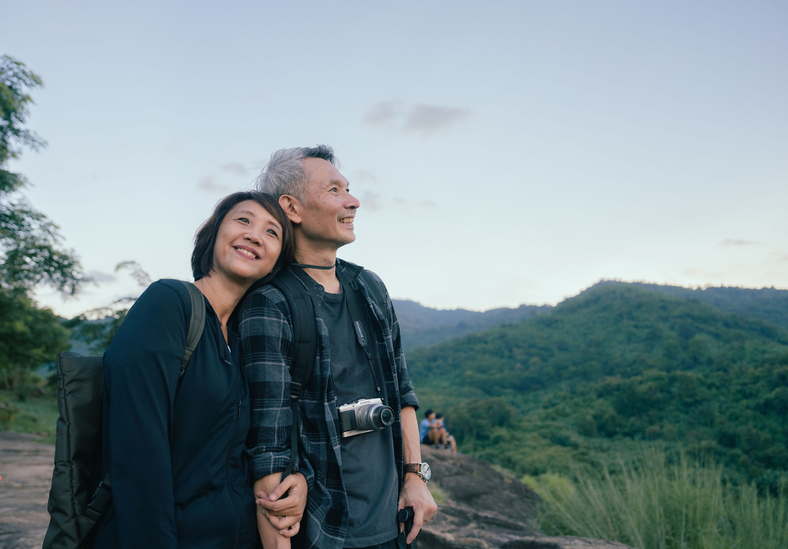 Happy Asian couple hikers on top of the mountain enjoying sunset over the tropical valley.