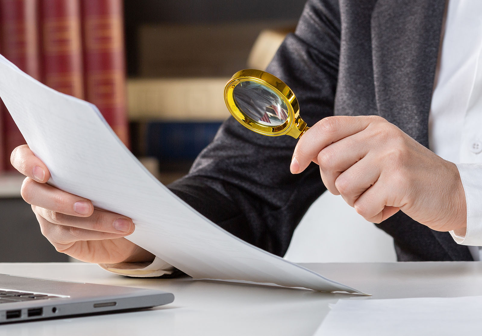 Female hands with a golden loupe and a papers at the desktop next to open laptop, close up, selected focus.