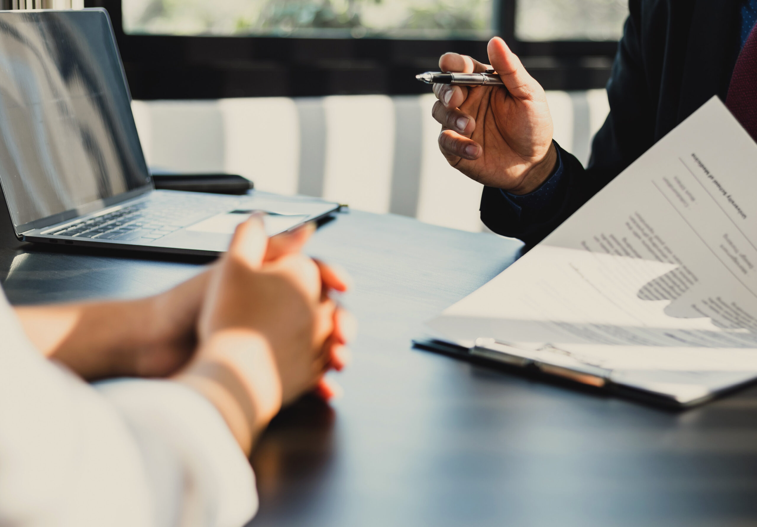Financial advisor meeting with a client at a modern office table. Advisor holds a pen and reviews a printed financial report with charts and graphs, while the client listens with clasped hands. An open laptop is on the table, sunlight streaming through large windows.