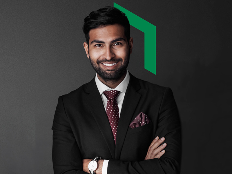 Smiling man in a dark suit and burgundy tie with a matching pocket square, standing with arms crossed against a dark background with a green geometric shape behind him.